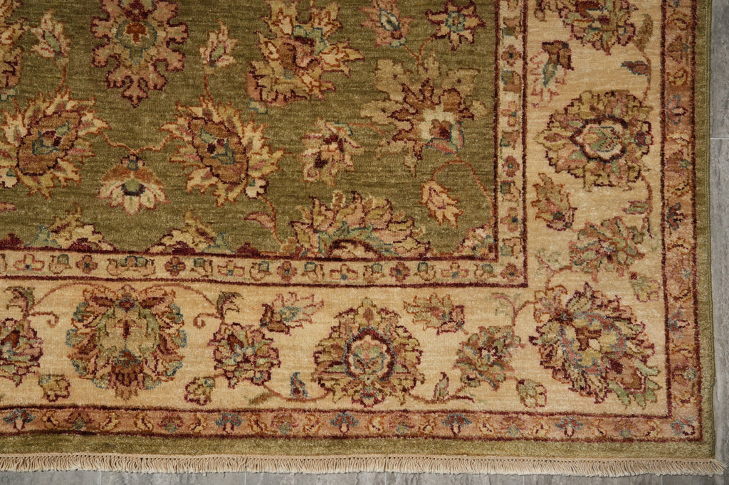 A close-up of an ornate traditional area rug showing its corner and border details. The rug features a muted olive green field with a beige inner border and a wide cream border decorated with floral motifs in brown, rust, and teal.