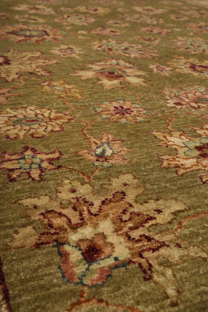 A close-up view of a traditional patterned area rug with a muted olive green base, featuring large floral motifs in beige, brown, and dusty pink with small accents of blue and red.