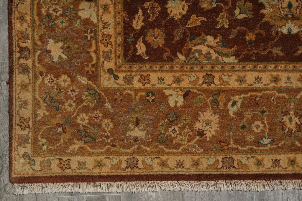 A close-up of an ornate area rug corner showing a dark brown field, beige and gold floral motifs, and a patterned border with fringe along the bottom edge.