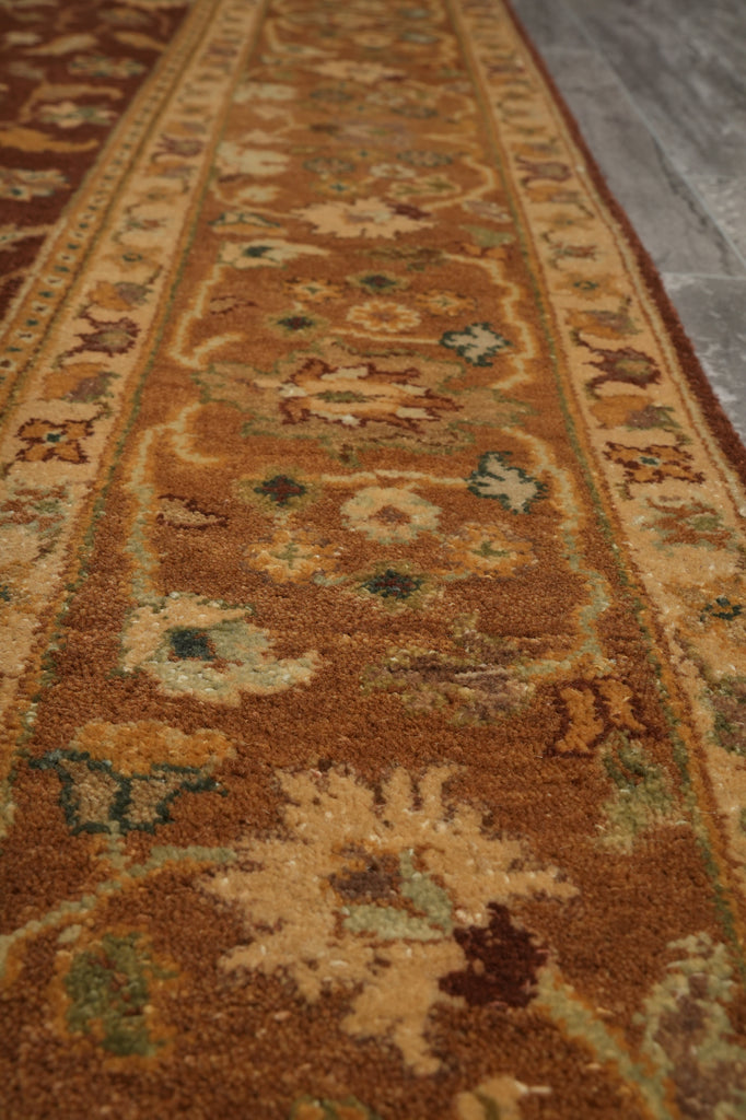 A close-up view of a traditional patterned area rug in warm brown, beige, and muted green tones, shown on a gray floor.