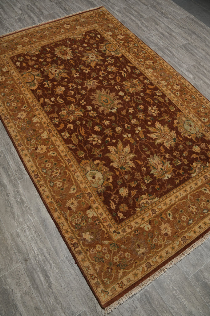 A rectangular traditional area rug with an intricate floral and vine pattern, shown from an elevated angle on a gray wood-look floor. The rug features a dark brown center field, a golden-brown border, and small light accents.