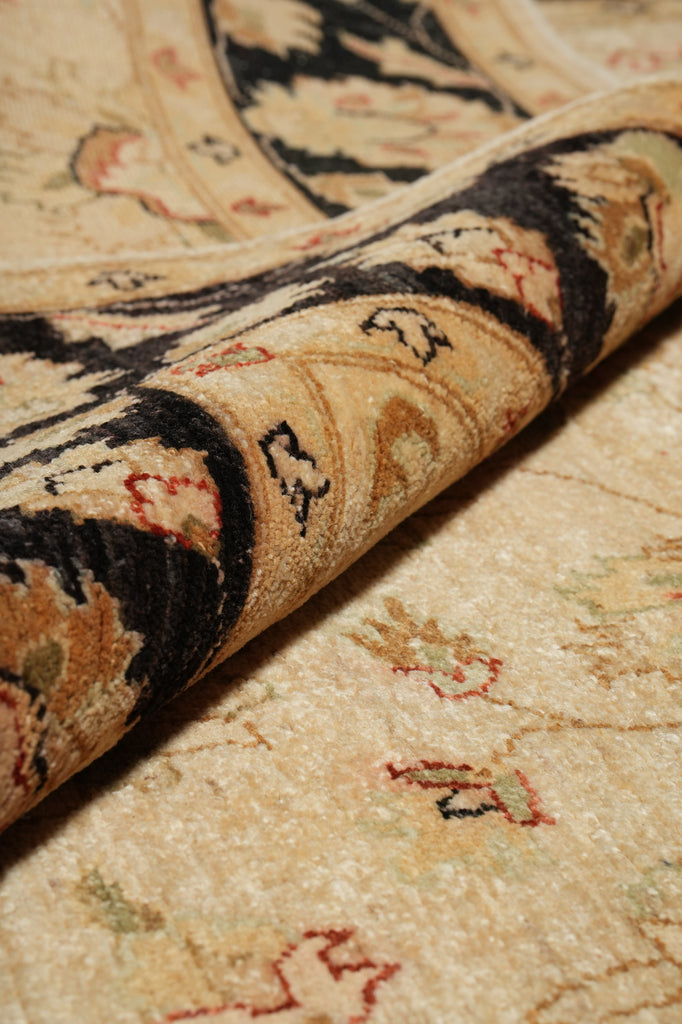 A close-up of a traditional patterned area rug with a rolled section showing the detailed pile and design. The rug features a beige base with black, brown, and red accents in an ornate floral and geometric motif.