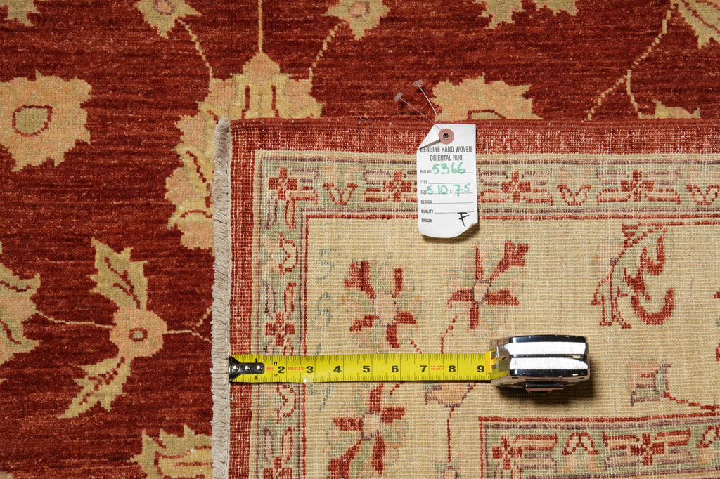 A close-up of a patterned area rug corner with a yellow tape measure and a white tag placed on top, showing the rug's red and beige floral design.