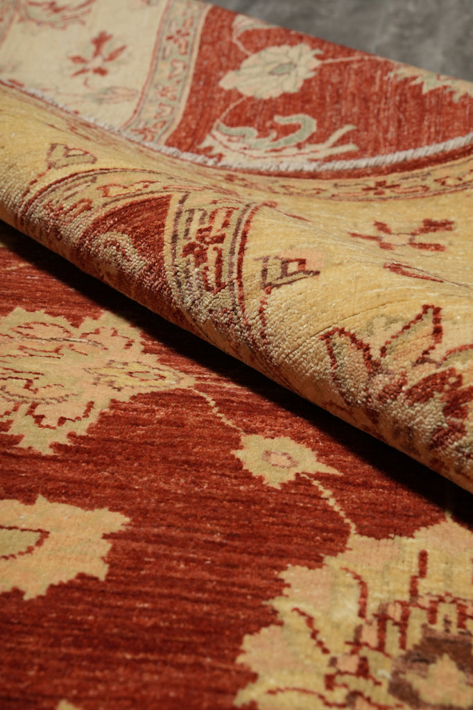 A close-up of a patterned area rug with a folded corner showing its texture and reverse side. The design features a warm rust red and beige floral motif.