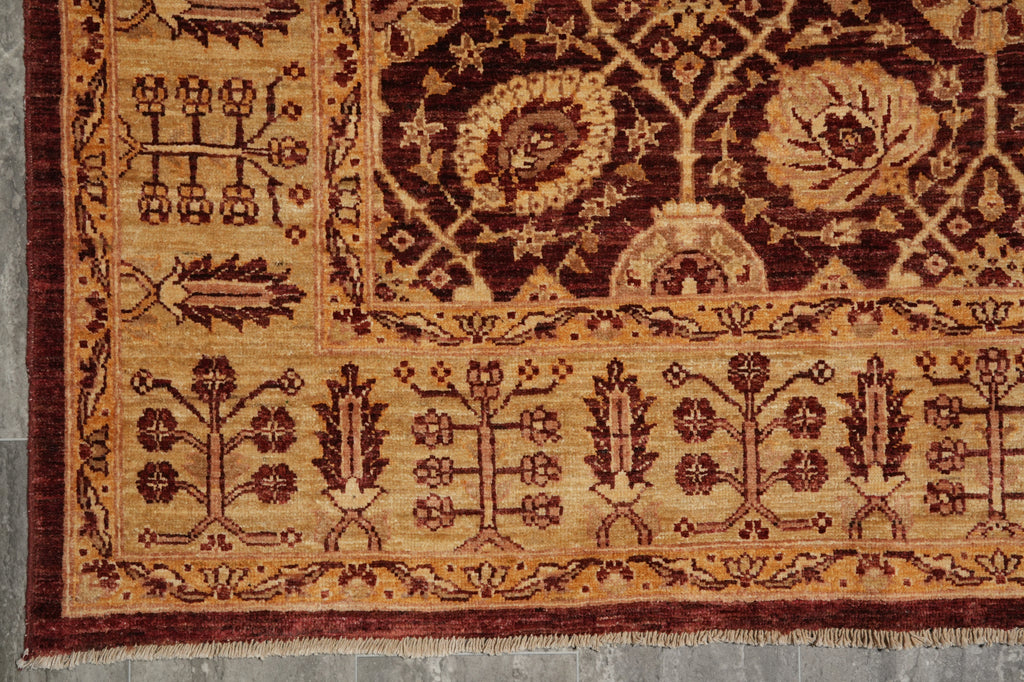 A close-up of an ornate traditional rug corner showing a beige and dark red pattern with floral motifs and a fringed edge on a gray floor.