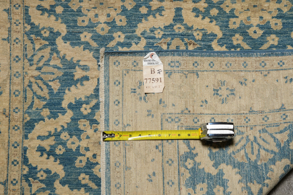 A close-up of a patterned area rug with a blue and beige floral design, shown with a yellow tape measure and a small inventory tag on top.