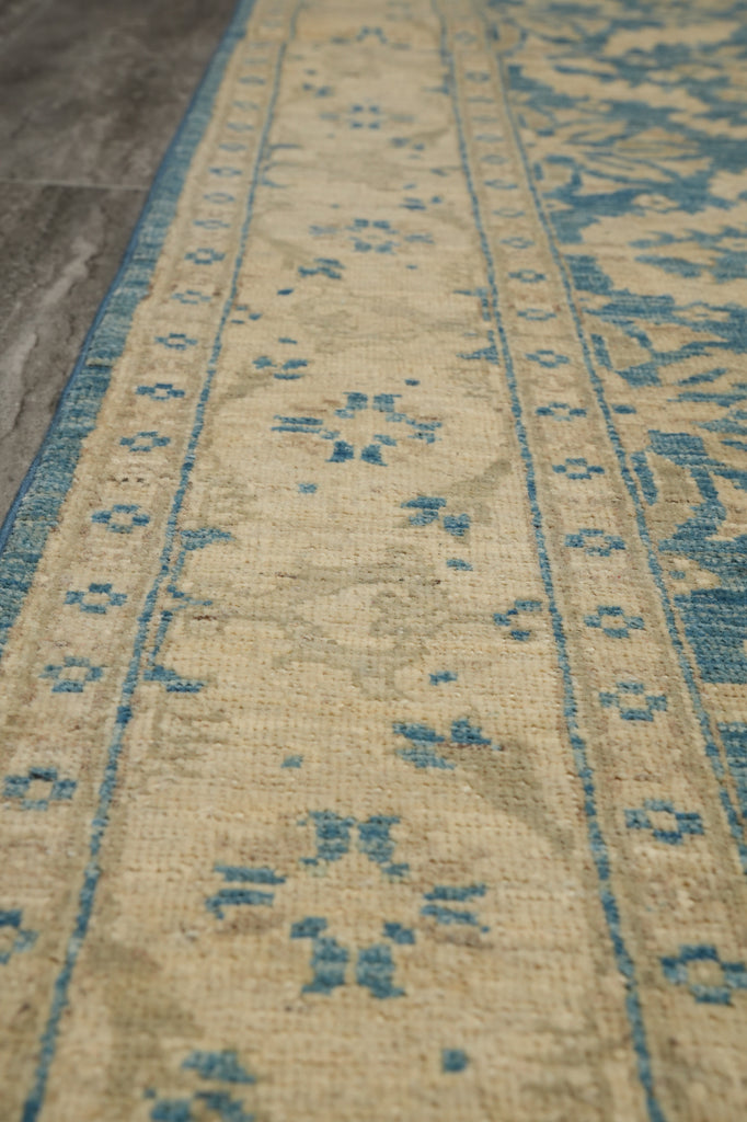 A close-up view of a traditional area rug with a beige base and blue distressed patterns, laid out on a gray tiled floor.