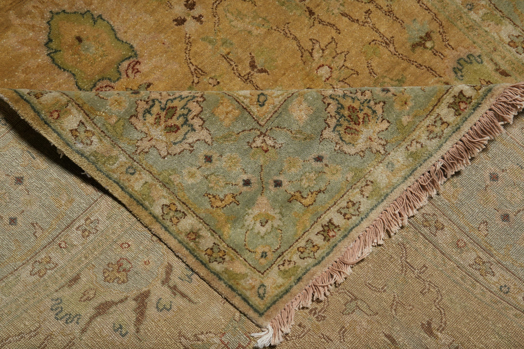 A close-up of a traditional patterned rug corner with fringe, showing a muted sage green border and floral motifs in beige, tan, and dusty pink tones over a warm brown field.
