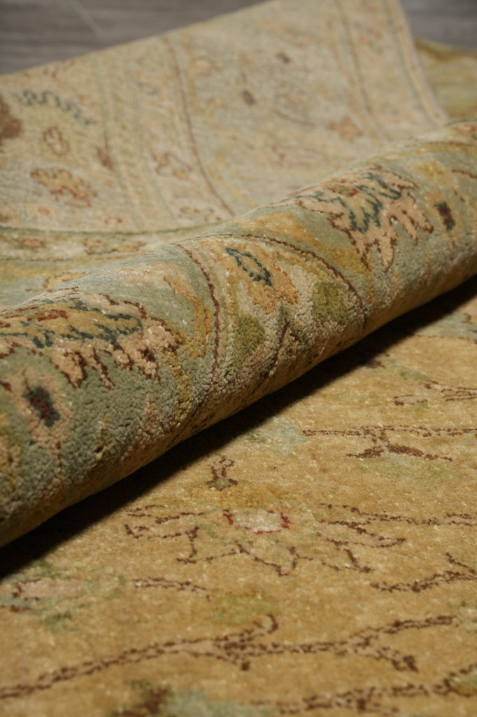 A close-up of a rolled section of an area rug showing its detailed vintage-style floral and botanical pattern in muted beige, sage green, and brown tones.