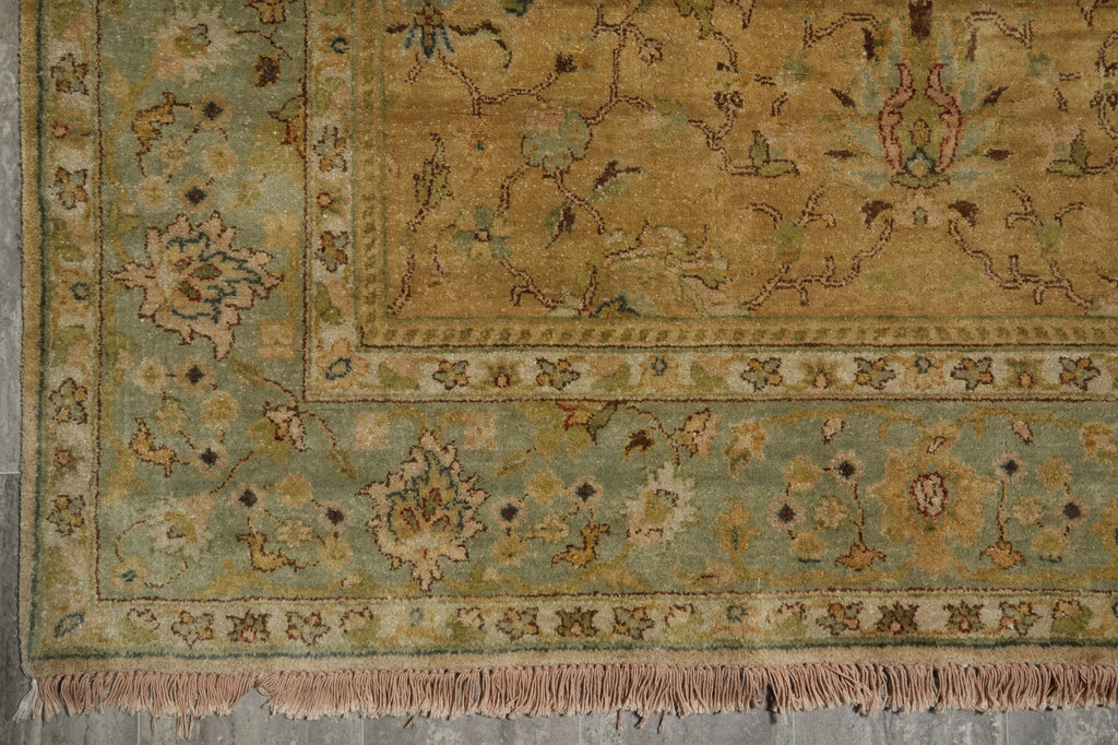 A close-up of an ornate area rug showing its corner, border, and fringe. The rug features a muted sage green and ochre yellow base with intricate floral motifs and traditional patterns.