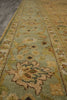 Load image into Gallery viewer, A close-up view of a traditional area rug with a light sage green field, beige and ochre floral motifs, and a patterned border, shown on a dark gray floor.
