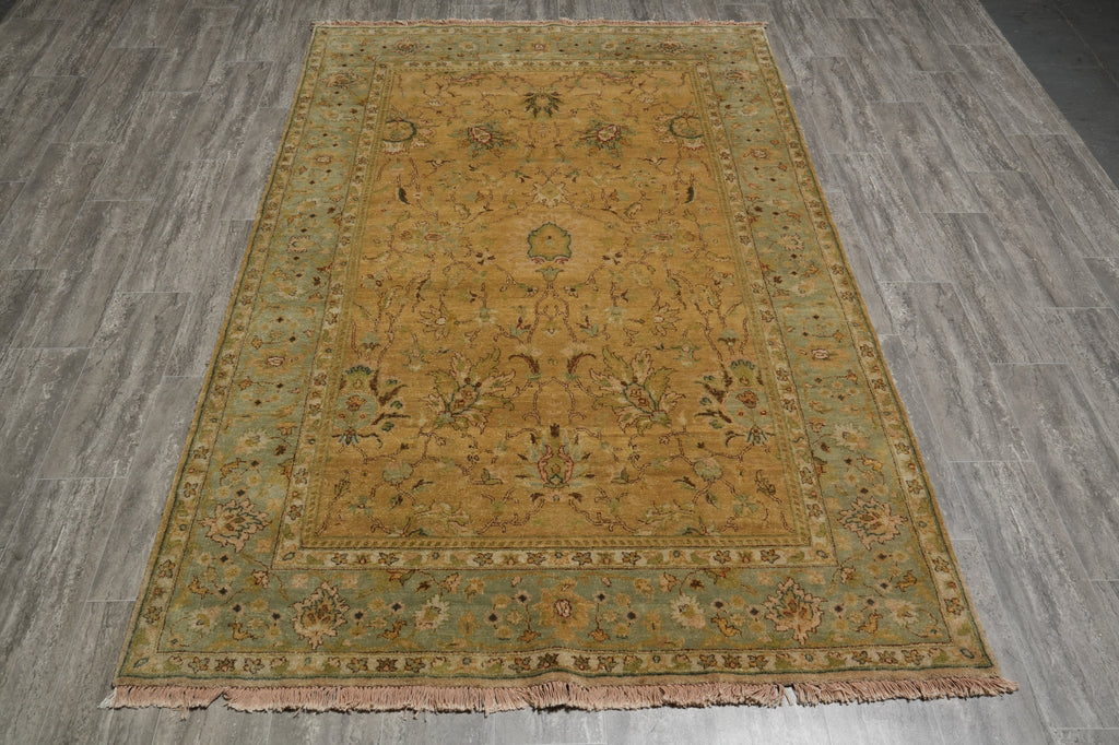 A rectangular vintage-style area rug with a central medallion and floral motifs, laid flat on a gray wood-look floor. The rug features a warm mustard-yellow center, a muted sage-green border, and dusty pink fringe.