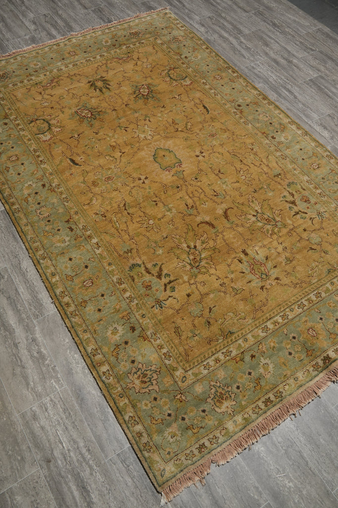 A rectangular area rug with a traditional floral pattern, featuring a mustard yellow center and a muted sage green border, laid out on a gray wood-look floor.