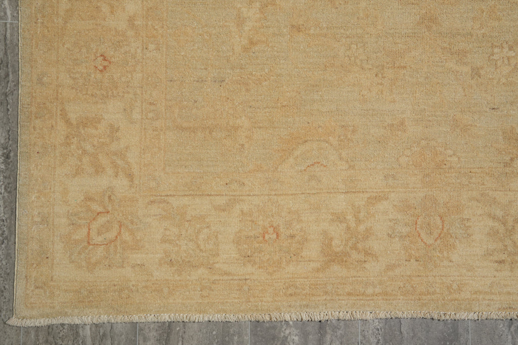 A close-up of a light beige area rug with a subtle faded floral pattern and fringed edge, laid on a gray tiled floor.