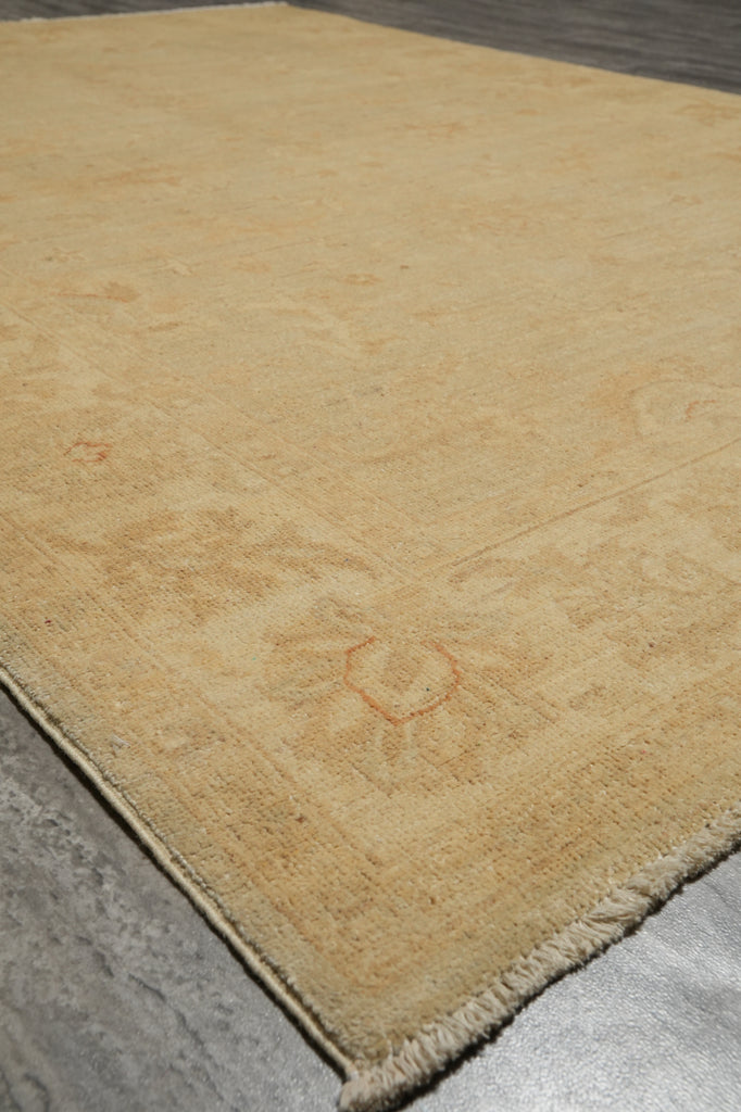A close-up of a light beige area rug with subtle faded floral patterns and fringed edges, laid out on a gray floor.