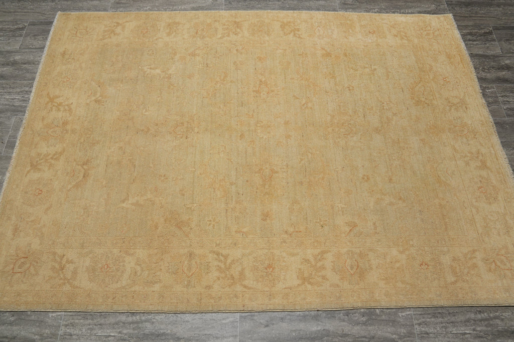 A large rectangular area rug in a warm sandy beige color with a subtle faded traditional floral pattern, laid flat on a dark gray tiled floor.