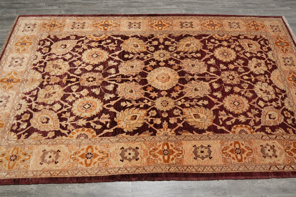 A rectangular traditional area rug with an intricate floral and geometric pattern, featuring a deep burgundy center field, warm beige and cream medallions, and a wide decorative border, laid out on a gray tiled floor.