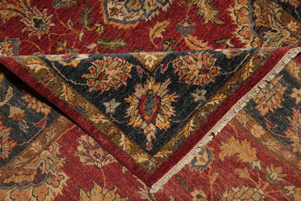 A close-up of an ornate patterned rug showing a folded corner that reveals a dark navy blue field with floral motifs in gold, red, and beige, layered over a larger red and gold patterned section.