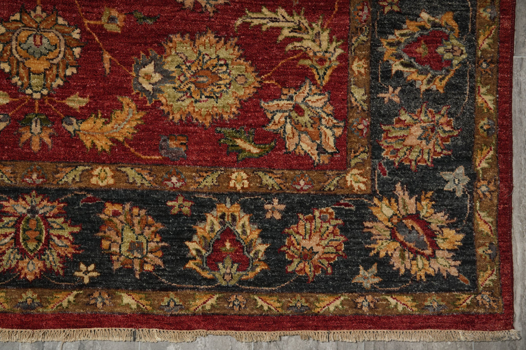 A close-up of an ornate area rug showing a deep red field with a dark charcoal-gray border and intricate floral motifs in beige, gold, and muted green.
