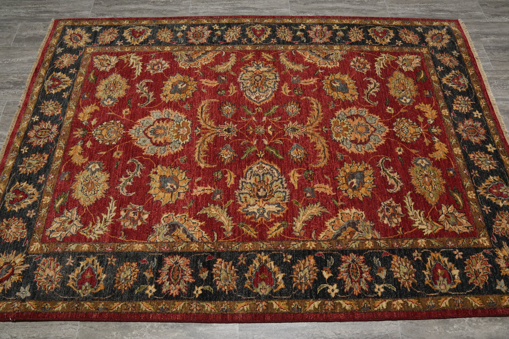 A traditional oriental area rug with a deep red field, featuring intricate floral and medallion motifs in gold, beige, and blue, surrounded by a dark navy border with gold detailing, laid out on a gray tiled floor.