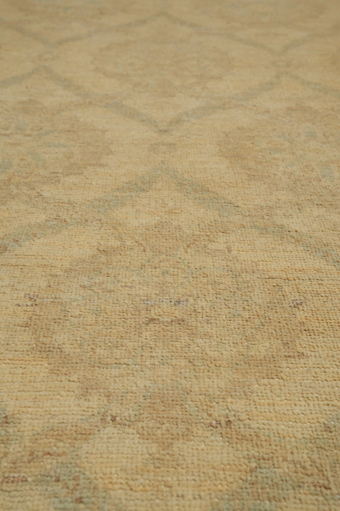 A close-up view of a textured area rug with a faded geometric diamond pattern in warm beige and light tan tones.