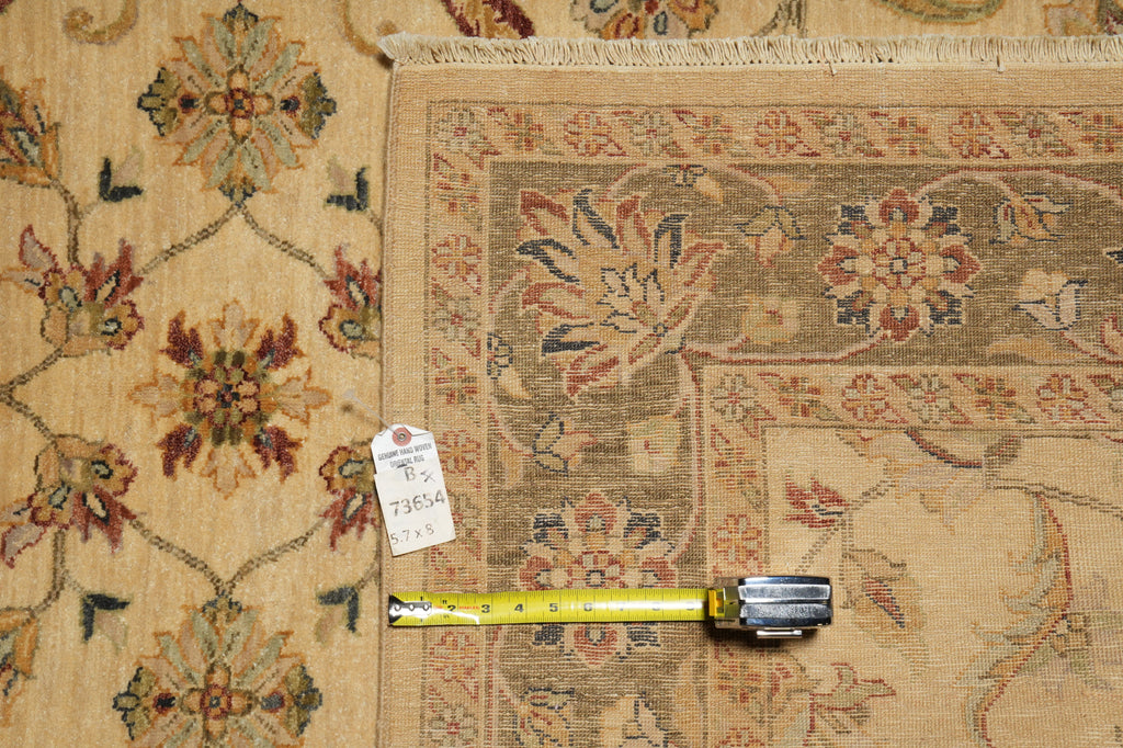 A close-up of a patterned area rug in beige, brown, and muted red tones, shown with a yellow tape measure and a small white tag on a matching rug surface.