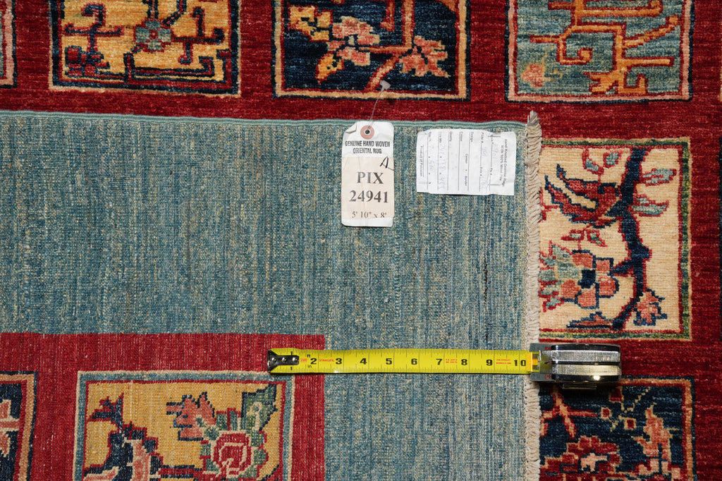 A close-up of a vintage patterned area rug showing a light blue-gray central field bordered by red and navy sections with floral motifs. A yellow tape measure is laid horizontally across the rug to indicate scale, with a white inventory tag attached near the top.