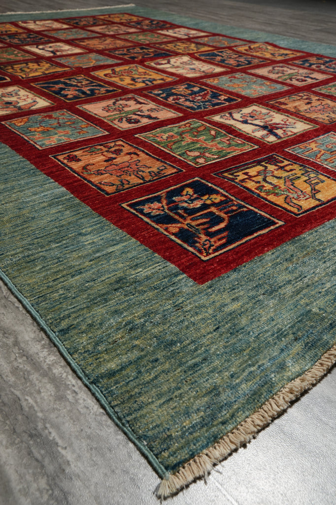 A traditional multi-panel area rug with a textured teal-green border and a central grid of colorful square motifs in red, blue, orange, and beige, laid out on a gray concrete floor.
