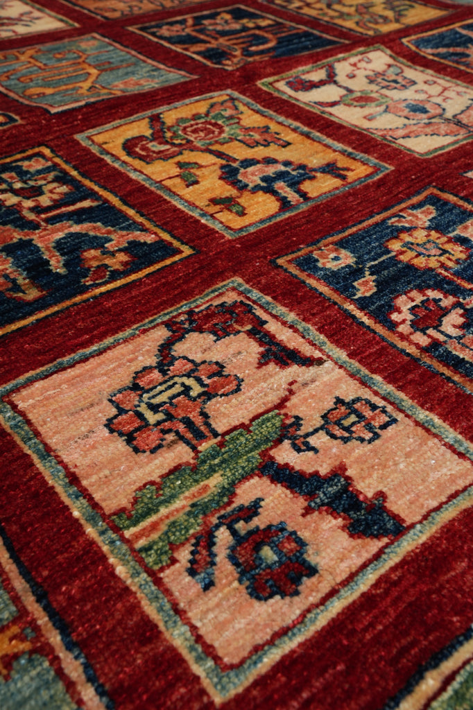 A close-up of a traditional patterned area rug with a deep red border and multiple square panels in beige, orange, and navy blue featuring floral and geometric motifs.