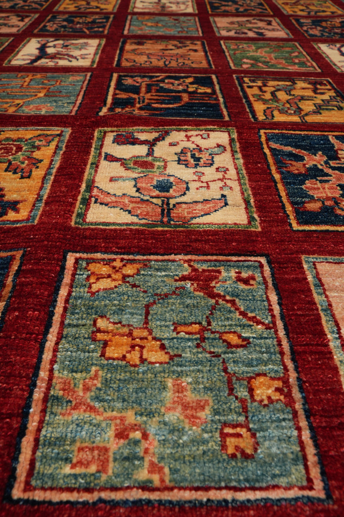A close-up view of a traditional hand-knotted area rug with a grid pattern of square panels. The rug features a deep red border framing panels with floral and geometric motifs in muted shades of blue, green, beige, and orange.