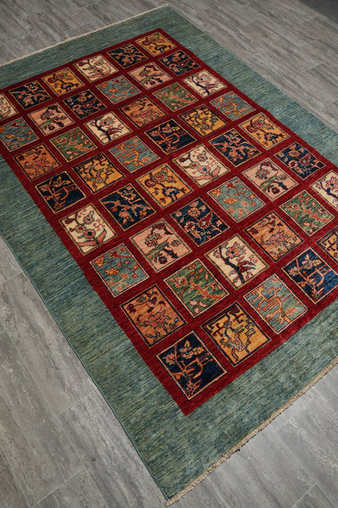 A rectangular area rug with a teal-green textured border and a central grid of square panels in red, orange, beige, and navy, shown laid out on a gray wood-look floor.