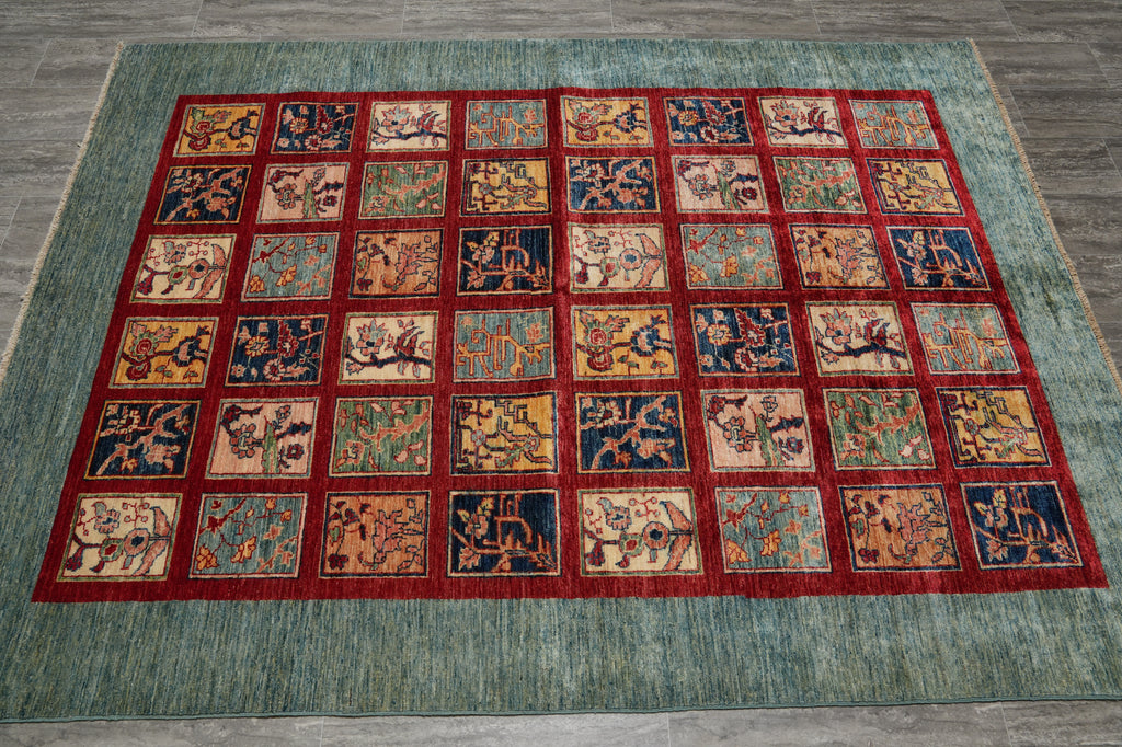 A large rectangular area rug with a teal-blue border and a grid of small multicolored medallion panels, photographed from above on a gray wood floor.
