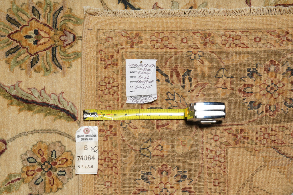 A close-up of a patterned area rug in beige and brown tones, laid flat with a yellow tape measure and two inventory tags placed on top to show dimensions and condition.