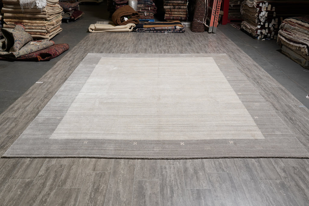 A large rectangular area rug with a light beige center and a textured taupe-gray border, displayed on a gray wood-look floor in a carpet showroom.