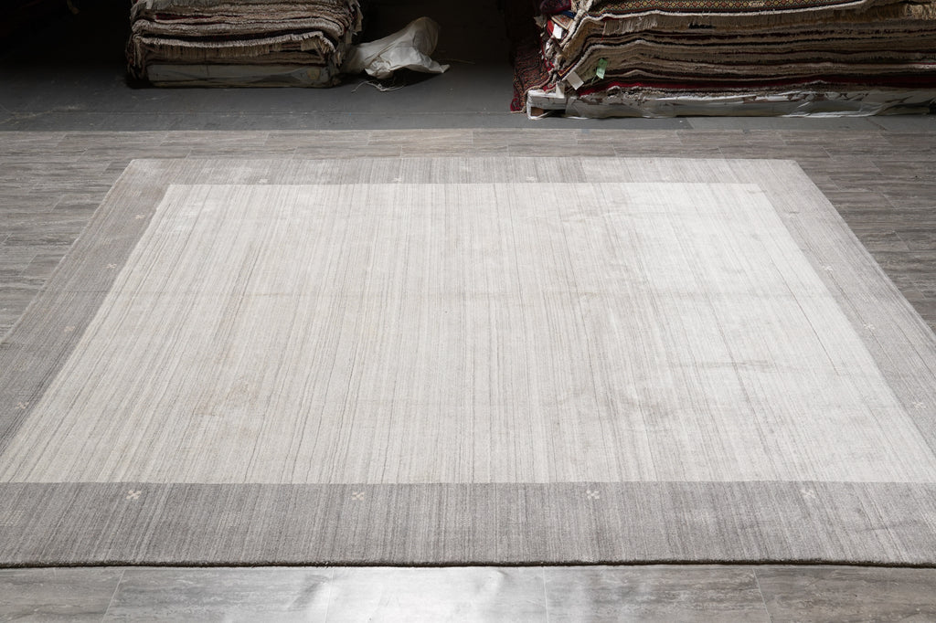 A large rectangular area rug with a light beige center and a medium gray border, laid out on a gray tiled floor in what appears to be a showroom or warehouse.