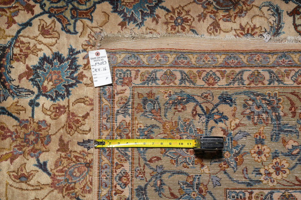 An ornate traditional area rug with a beige, blue, and rust floral pattern, shown with a yellow tape measure and a white tag on a flat surface.