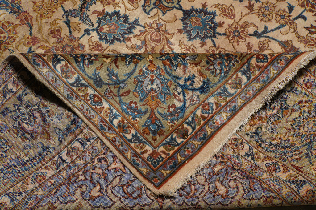 A folded traditional oriental rug with an intricate floral and vine pattern in beige, blue, brown, and rust tones, showing the corner design and fringed edge.