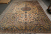 Load image into Gallery viewer, A large traditional oriental area rug with an intricate floral pattern in beige, blue, and brown tones, shown laid out on a gray floor with a person's legs partially visible on the right edge.
