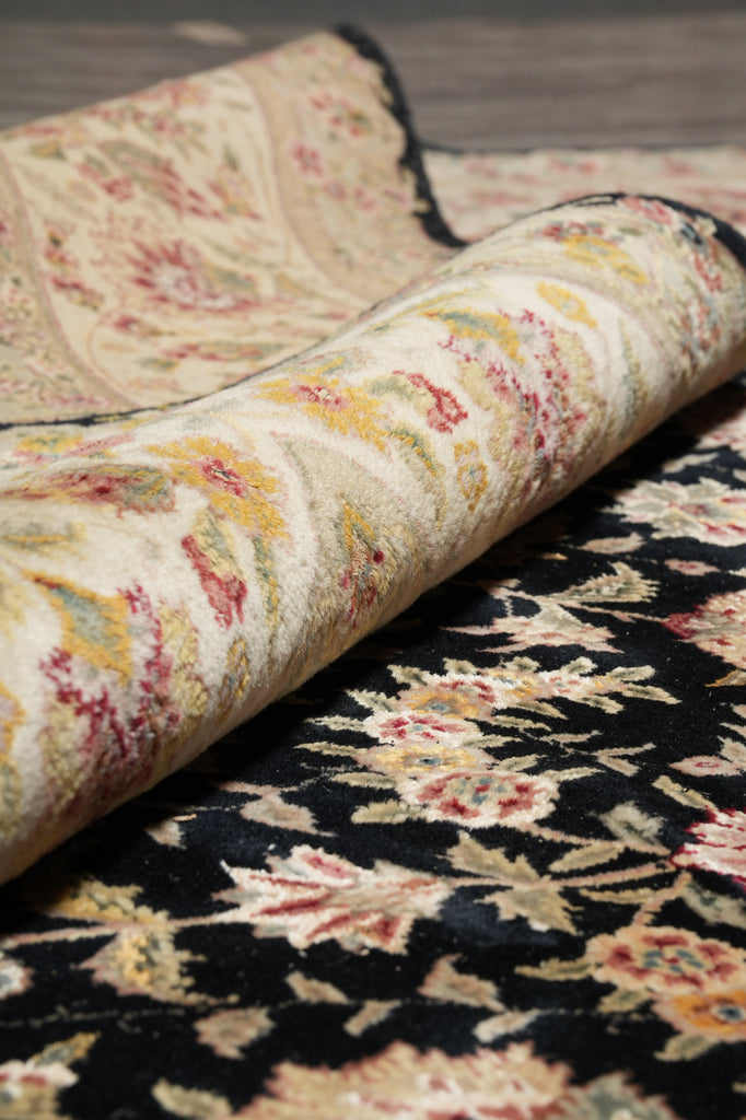 A close-up of a folded patterned rug showing two distinct sections: one with a black background and cream floral motifs, and another with a beige background and multicolor floral/paisley patterns.