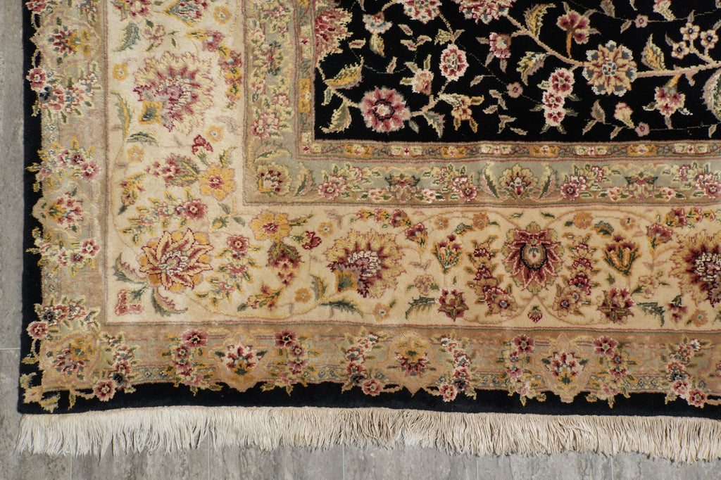 A close-up of an ornate area rug corner showing a cream floral field, detailed borders in beige and burgundy, a black central field with floral motifs, and cream fringe at the bottom.