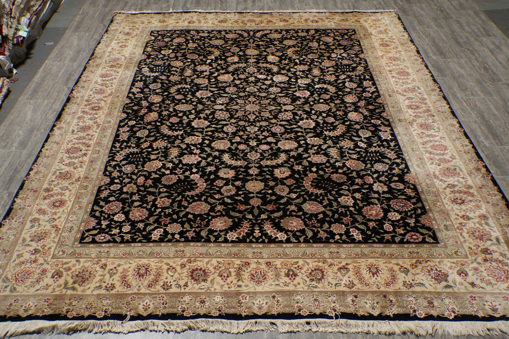 A rectangular area rug with a black central field covered in dense floral motifs, surrounded by a wide beige border with matching floral patterns, photographed from above on a gray floor.