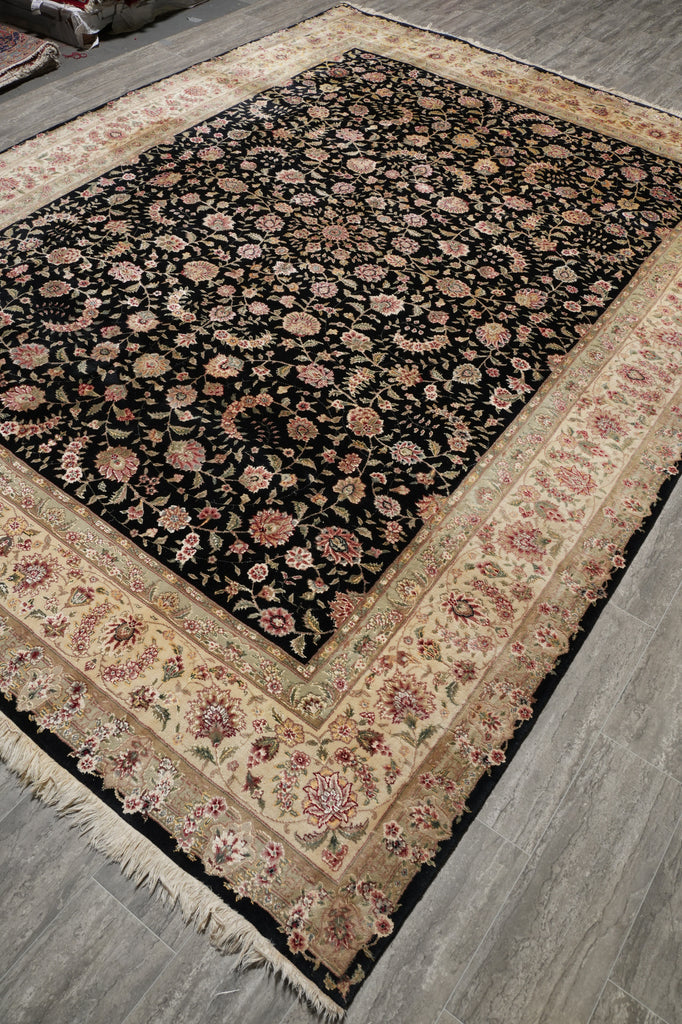 A traditional oriental area rug with a black field, beige borders, and pink floral motifs, shown laid out on a gray wood floor.