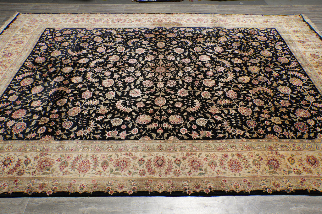 A rectangular area rug with an intricate floral pattern, featuring a black center field and a beige border, laid out on a gray tiled floor.