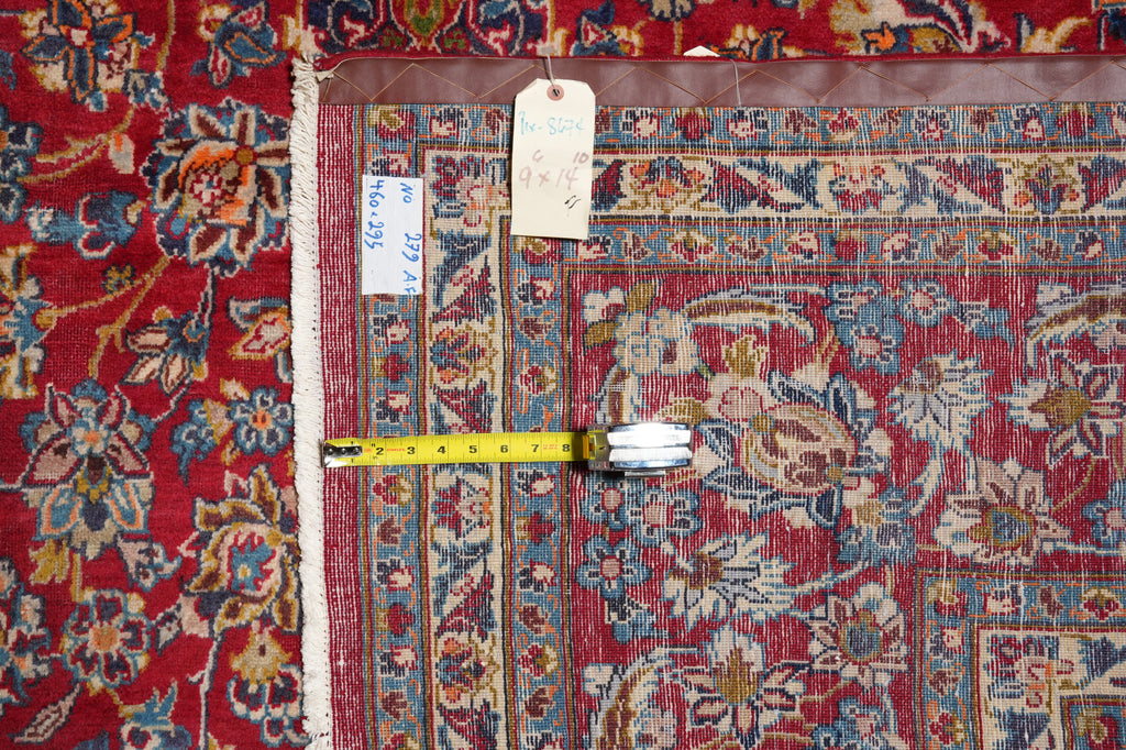 A close-up of a patterned area rug with a red field, blue and beige floral motifs, and a brown border, shown with a yellow tape measure and handwritten tags for size reference.