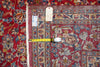 Load image into Gallery viewer, A close-up of a patterned area rug with a red field, blue and beige floral motifs, and a brown border, shown with a yellow tape measure and handwritten tags for size reference.