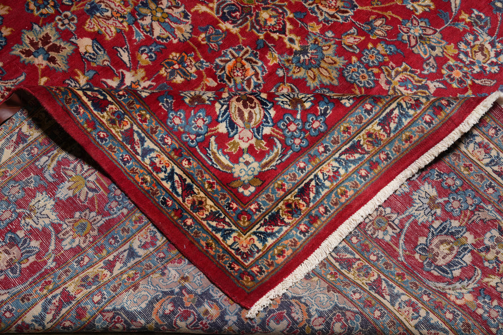 A close-up of a traditional oriental rug with a deep red field, intricate floral motifs in blue, beige, and gold, and a decorative border with white fringe.