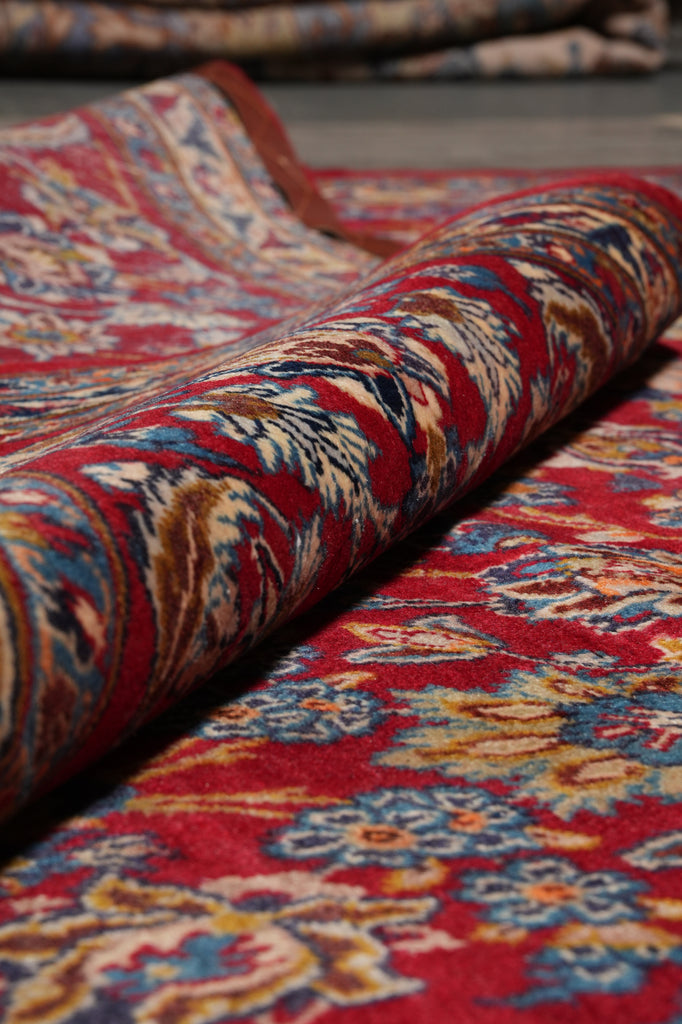 A close-up of a folded patterned rug showing a traditional floral design in red, blue, beige, and brown tones.