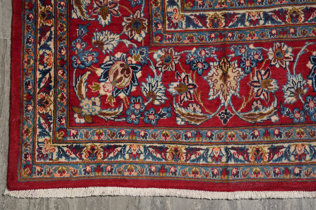 A close-up of a traditional patterned rug corner, showing a deep red field with ornate floral motifs and a detailed multi-colored border in blue, beige, and gold, finished with white fringe.