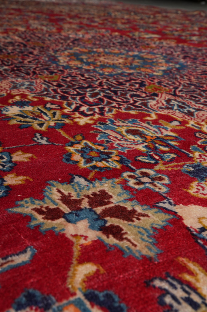 A close-up view of a traditional patterned area rug with a vibrant red field, featuring intricate floral motifs in blue, beige, and brown.