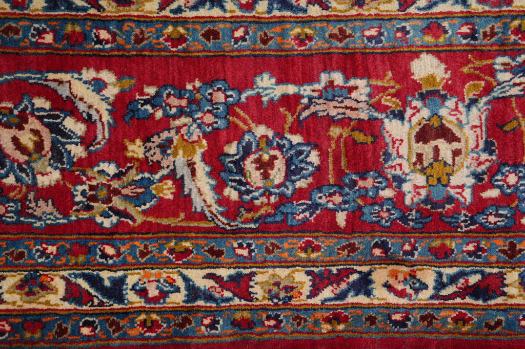 A close-up of a traditional patterned rug featuring a vibrant red field with intricate floral motifs in blue, beige, and gold, bordered by detailed floral bands.